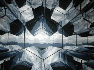 ID: A landscape phot of a series of honeycomb-shaped mirrors, reflecting the view of a mountain range in the distance. End ID.